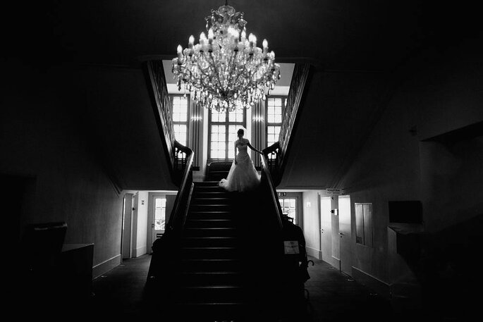 Eine Braut steht am Fuße einer Treppe. Über ihr thront ein Leuchter.