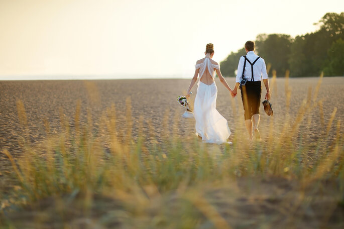 Un couple de jeunes mariés se balade main dans la main, dans un champ. La mariée porte une longue robe blanche et élégante. Le marié porte une chemise blanche et un bas de pantalon noir. 
