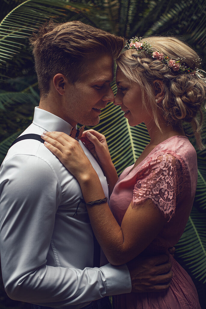 Kurz vor dem Kuss: Ein Brautpaar liegt sich lächelnd in den Armen, ihre Nasen berühren sich bereits. Im Hintergrund ist ein grüner Garten zu sehen.