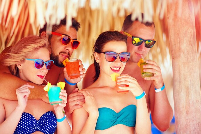 group of happy friends having fun on tropical beach, drinking colorful cocktails