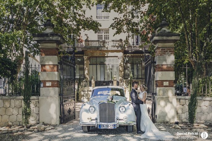 Deux mariés enlacés à côté d'une voiture de collection blanche, devant le portail du Château la Beaumetane