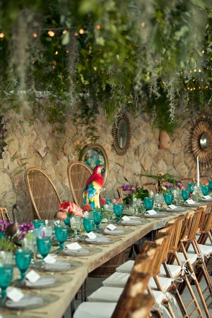 Mesa de convidados com taças turquesa e decoração tropical com flores e arara de porcelana.