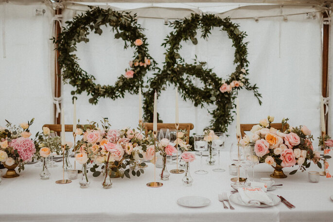 Table décorée dans les tons pastels avec des couronnes de végétaux en arrière plans, des bougies et des bouquets de fleurs pastel 