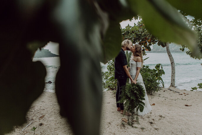 Das Brautpaar küsst sich am Strand, vor der Linse sind grüne Blätter zu sehen.