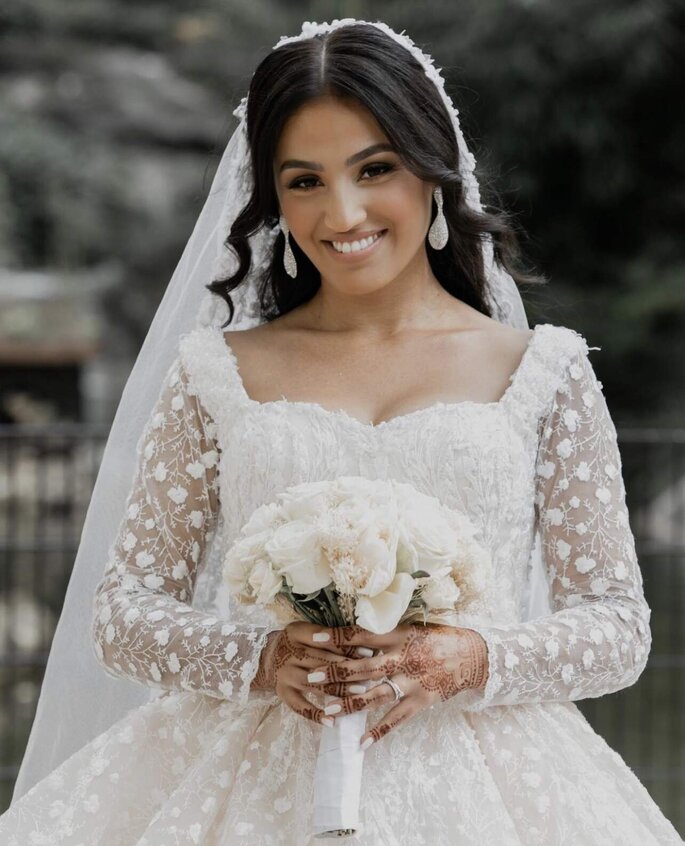 Une mariée tient un bouquet de fleurs blanches, elle porte un voile et une robe princesse avec des manches et des jeux de transparence