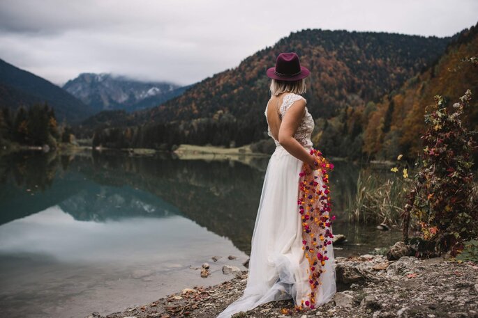 une mariée porte une robe de mariée dos nu avec un haut en dentelle, un voile avec des fleurs cousues et un chapeau violet 