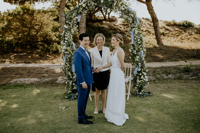 une affinate de cérémonie rient avec un couple de mariée, ils sont dans un parc, devant une arche de fleurs 