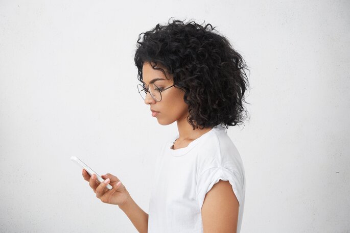 Sobre um fundo na cor branca temos uma mulher negra de cabelos curtos utilizando um aparelho celular.