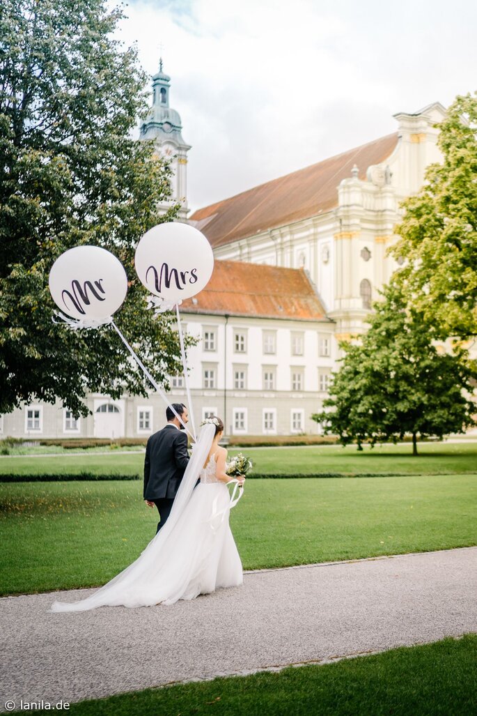 Brautpaar mit Mr & Mrs Luftballons auf einer Wiese vor einer Kirche