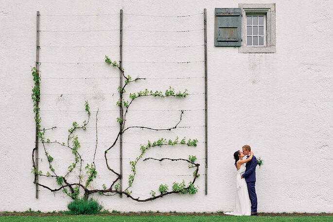 Ein Brautpaar küsst sich vor einer weißen Wand, an der Efeu rankt.