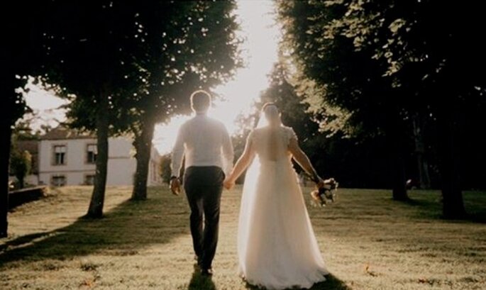 Un couple de mariés qui se tient la main avance dans une allée bordée par des arbres.
