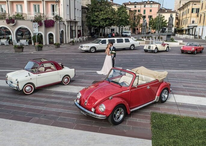 sposi al centro di un piazzale, varie auto d'epoca rosse bianche intorno